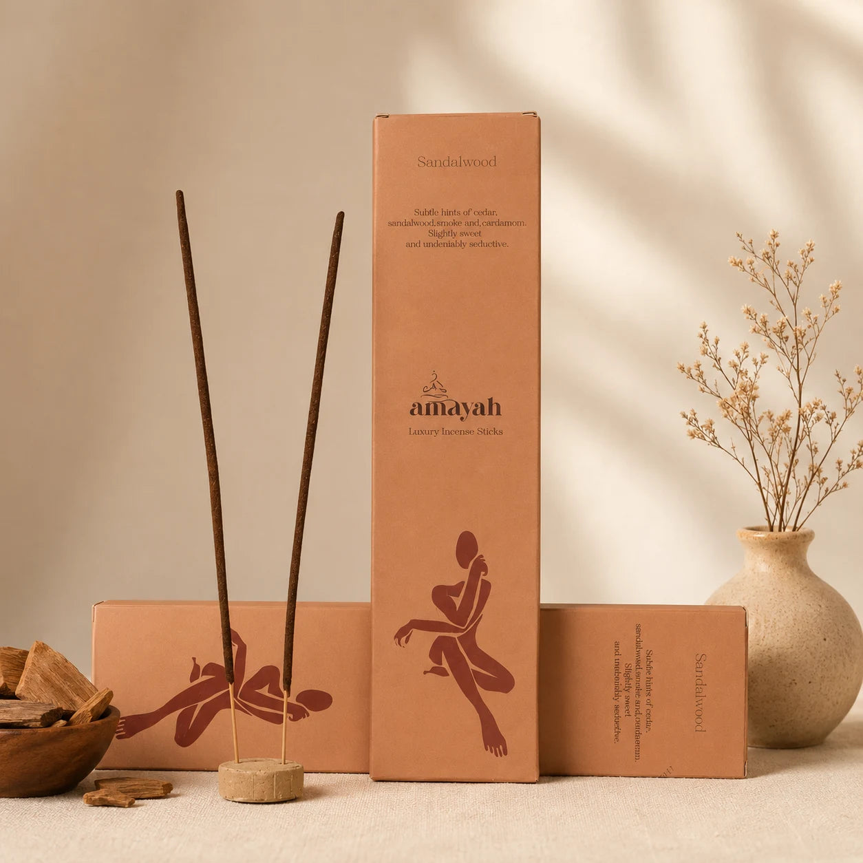Amayah chandan agarbatti and packaging boxes arranged on a neutral surface with a beige wall backdrop, plus a small vase of dried stems and a bowl of incense pieces. The product packaging is tan with dark brown printed graphics and text, and the incense sticks are dark brown.